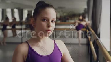 穿着紧身衣的黑发小女孩站在芭蕾舞课上看着相机的特写肖像。 其他学生